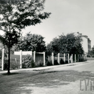 brukowana ulica, wzdłuż niej ogrodzenie z metalowej siatki, za nim ogródki