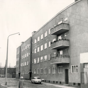 ulica gruntowa, przy niej czterokondygnacyjny blok mieszkalny z jednym rzędem balkonów na trzech kondygnacjach, przy budynku niski, murowany pawilon handlowy, przed blokiem zaparkowany samochód osobowy, z lewej niewielki fragment ceglanego budynku mieszkalnego