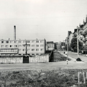 na pierwszym planie gruntowa droga, z lewej kamienne ogrodzenie z wkomponowanym krzyżem, za nim kilkukondygnacyjny budynek, z prawej brukowana ulica, po jej prawej stronie wysokie kamienice