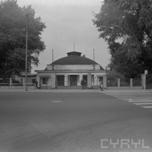budynek z rozległą kopułą otoczony płotem, na chodniku przed budynkiem przechadzają się przechodnie, obok rosną drzewa