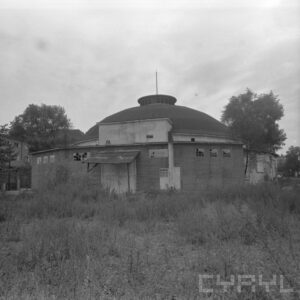 zniszczony budynek z kopułą otoczony krzewami; w głębi z boku widoczne budynki mieszkalne