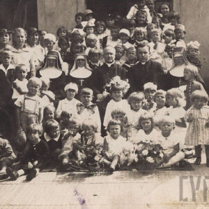 duża grupa mniejszych i większych dzieci, z przodu siedzą, w dalszych rzędach stoją, między nimi kilkoro dorosłych, a wśród nich trzy siostry zakonne w kornetach