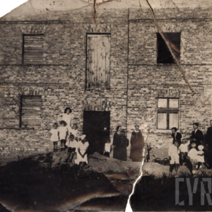 dorośli z licznymi małymi dziećmi stoją przed nowo wybudowanym, nieotynkowanym, piętrowym domem z cegły z zabitymi deskami oknami po lewej stronie