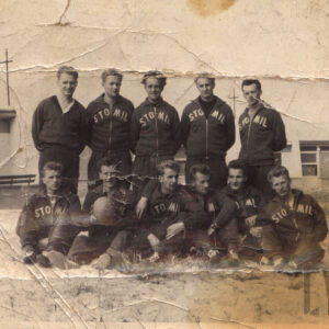 grupa młodych mężczyzn w dresach klubowych, sześciu z nich siedzi na trawie, pozostali stoją nad nimi. w głębi niezidentyfikowany budynek z wejściem i oknami na parterze