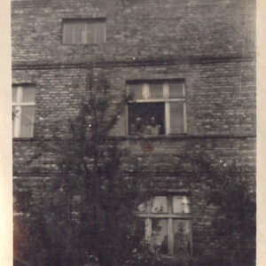 fragment ceglanej, nieotynkowanej ściany piętrowego domu z poddaszem, po dwa okna na parterze i na piętrze, jedno w partii poddasza, w oknie na pierwszym piętrze dwoje ludzi