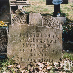 Uszkodzony kamienny nagrobek z inskrypcją. Złamana górna część z krzyżem. Ślady po pociskach. Za nim inne nagrobki.