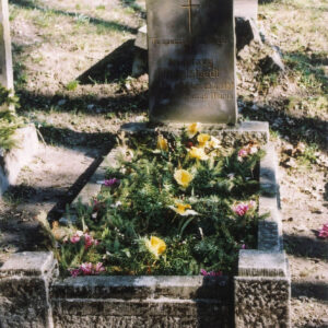 Nagrobek w kształcie pionowej, prostokątnej steli z napisami i pionowym krzyżem łacińskim. Wąskie obmurowanie z parą narożnych słupków wypełnione gałązkami świerku i kwiatami. Za nim inne nagrobki.  