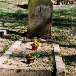 Nagrobek otoczony niskim obmurowaniem. Na nim trzy niewielkie znicze pośrodku. Pionowa prostokątna stela z napisami i krzyżem łacińskim. Za nim inne nagrobki i drzewa.   