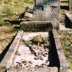 Nagrobek otoczony murkiem. Płyta nagrobna z otworem w kształcie rombu pośrodku. Stela z napisami i wykutym krzyżem odspojona od obmurowania. Nad inskrypcją ceramiczny medalion. W głębi fragmenty innych nagrobków.