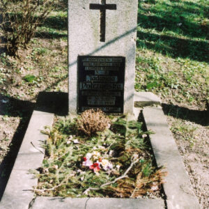 Nagrobek otoczony szerokim murkiem z wyłożonymi gałązkami świerku. Prostokątna stela z łacińskim krzyżem z czarnego szkła. Tablica epitafijna z czarnego szkła z napisami. Z tyłu inne nagrobki, krzew i pnie drzew.