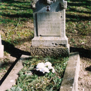 Nagrobek otoczony murkiem z wyłożonymi w środku gałązkami świerku i białymi sztucznymi kwiatami. Stela z tablicą epitafijną, medalionem portretowym i płaskorzeźbą uskrzydlonej głowy anioła. Z tyłu teren porośnięty trawą i krzewami.