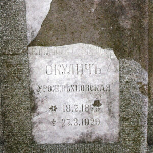 Uszkodzona biała, prostokątna tablica z napisami cyrylicą wtopiona w płytę nagrobną.