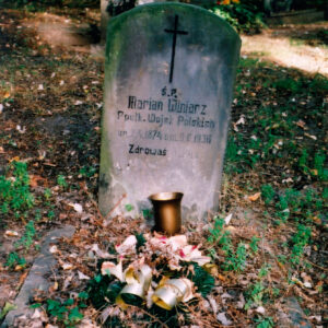 Stela z napisami i krzyżem łacińskim. Niskie obmurowanie, na którym stoją wiązanka i pusty wazon. Teren pokryty suchymi liśćmi. W głębi inne nagrobki. 