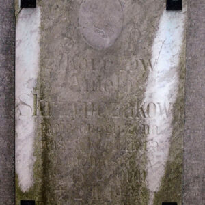 Prostokątna biała tablica z napisami i śladem po medalionie portretowym przytwierdzona w narożach czterema metalowymi nitami.