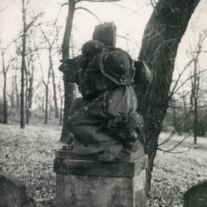 Pomnik mężczyzny obejmującego krzyż na postumencie. Ubrany w długą szatę, na plecach ma kapelusz. Z tyłu drzewa.