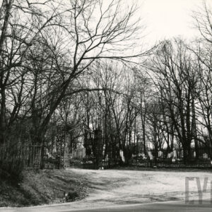 Metalowa brama dwuskrzydłowa i płot. Fragment drogi. Za płotem drzewa i kamienne nagrobki.