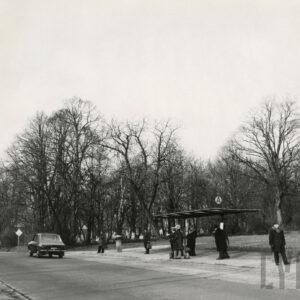 Przystanek autobusowy przy ulicy, po której jedzie samochód i autobus. Na przystanku stoją ludzie. Z tyłu za drzewami ukryty cmentarz.