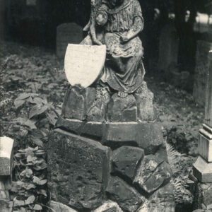 Figura anioła siedzącego na kamiennym kopczyku. Przy nim fragment owalnej tablicy z napisami. Z tyłu fragmenty innych nagrobków.