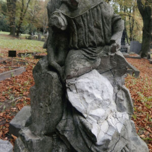 Zniszczona i zabrudzona figura anioła siedząca obok pnia drzewa. Brak głowy, odłamane ręce i duży fragment nóg. Z tyłu inne nagrobki.
