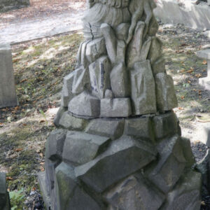 Tył zniszczonej figury anioła siedzącej obok pnia drzewa na kamiennym kopczyku. Z tyłu alejka i inne nagrobki.