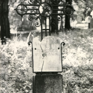 Żelazny krzyż na cokole z tablicą z napisami u podstawy. Ramiona zakończone kołami. Z tyłu drzewa i wysoka trawa.