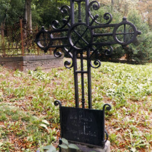 Żeliwny, ażurowy krzyż osadzony na kamiennym cokole z ramionami zakończonymi okręgami. Za nim żelazne ogrodzenie grobu, krzewy i drzewa.    