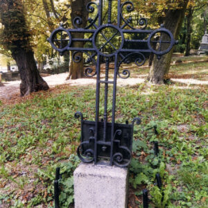 Tył żeliwnego krzyża na kamiennym postumencie. Za nim drzewa i fragmenty nagrobków. 