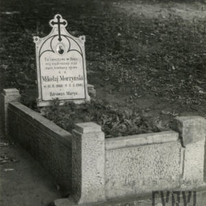 Obmurowany grób ze  skośnie ustawioną tablicą, na której szczycie jest krzyż łaciński, a pod nim medalion portretowy i napisy. Z tyłu teren pokryty suchymi liśćmi.