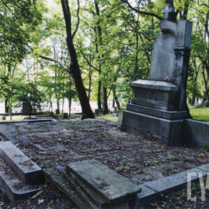 Duży grobowiec w kształcie ołtarza z krzyżem łacińskim na szczycie. Zniszczone obmurowanie mogiły i schody. Z tyłu drzewa. 