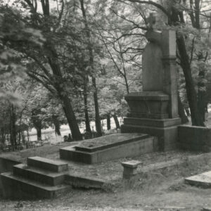 Duży, kamienny nagrobek w formie ołtarza z krzyżem na szczycie. Zniszczone obmurowanie i schody. Z tyłu drzewa i opłotowania nagrobków.