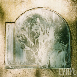 Zabrudzona plakieta z wizerunkiem lewego profilu mężczyzny wtopiona w płytę nagrobną.