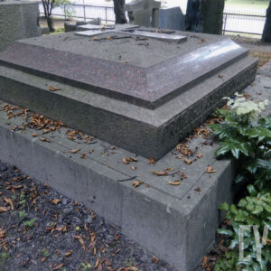 Duży nagrobek z poziomą płytą w kształcie kwadratu, na której wykuto krzyż łaciński. Na boku podstawy widnieje napis. Z tyłu ogrodzenie cmentarza, fragmenty innych nagrobków i drzew.