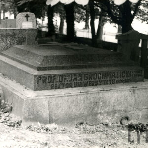 Duży nagrobek w kształcie poziomo leżącej płyty z krzyżem łacińskim. Na boku podstawy widnieje napis. Z tyłu drzewa, inne nagrobki i ogrodzenie.