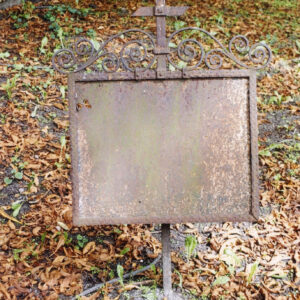 Pokryta rdzą tablica umocowana na pręcie zakończonym krzyżem łacińskim, ze ściętymi ukośnie zakończeniami ramion. Wokół suche liście.