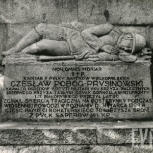 rzeźba przedstawiająca żołnierza w polowym mundurze z hełmem na głowie, leżącego w nurtach symbolizowanej falistymi liniami rzeki, obejmującego ręką drzewce sztandaru uwieńczone orłem