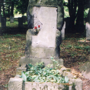 Nagrobek z wysokim obmurowaniem i pionową, wąską stelą. Na niej uszkodzona tablica epitafijna z krzyżem w górnej części. Za nagrobkiem drzewa i inne groby.