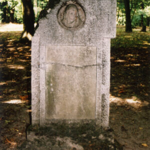 Pionowa prostokątna płyta nagrobna z odłamanym lewym, górnym narożem. W górnej części medalion z wizerunkiem Matki Boskiej, poniżej napisy. Z tyłu pnie drzew i zieleń.