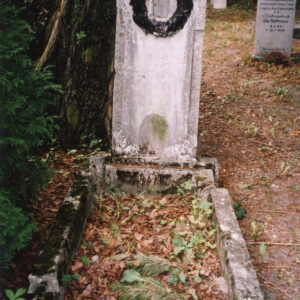 Zabrudzony nagrobek z obmurowaniem i wysoką stelą. W górnej części metalowy, laurowy wieniec ze śladem po plakiecie w środku. Wokół inne nagrobki i drzewa.