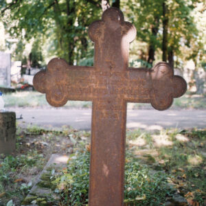 Pokryty rdzą krzyż łaciński z inskrypcją. Za nim inne nagrobki i drzewa.