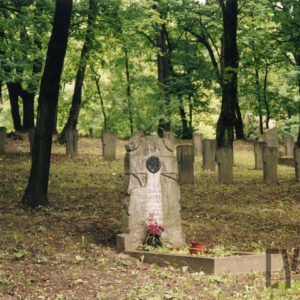 Nagrobek z pionową płytą nagrobną i plakietą z napisem Ave Maria. Za nim wiele innych nagrobków. Pomiędzy nimi drzewa.