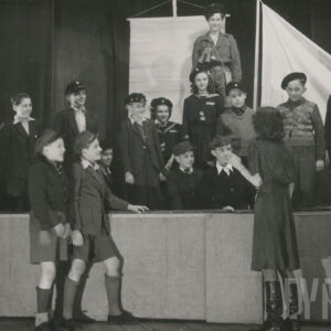 scena na statku, grupa młodych aktorów stoi obok siebie w różnych pozach, z lewej strony pochyla się dorosły aktor, przed statkiem stoi dwójka młodzieży obok nich odwrócona tyłem kobieta w sukni w kropki