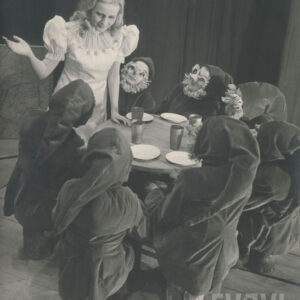 przy okrągłym stoliku siedzą na drewnianych pieńkach aktorzy w strojach krasnoludków, przy stole stoi kobieta w białej sukni, opiera się prawą ręką o blat, lewą rękę ma zgiętą w łokciu i podniesioną