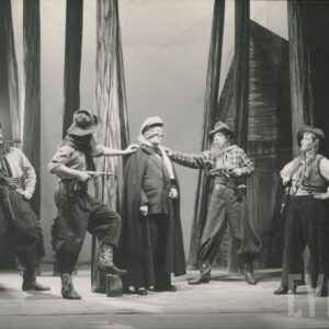 scena w lesie, pośrodku stoi mężczyzna w garniturze i pelerynie z opaską na oczach, wokół niego stoją aktorzy w koszulach w kratę, szerokich spodniach i pasach, w rękach trzymają rewolwery, obok stoi kobieta w szerokich spodniach, kamizelce i kapeluszu, ręce podpiera w pasie