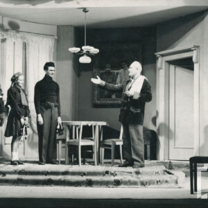 scena w pokoju, mężczyzna ubrany w spodnie w pasy i marynarkę wyciąga prawą rękę w kierunku stojących naprzeciw kobiety i dwóch mężczyzn, którzy trzymają łyżwy 