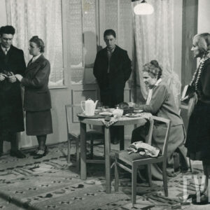 przy stole siedzi kobieta w koku, obok stoi dziewczyna z długimi warkoczami i książką, za stołem stoi mężczyzna trzymający ręce w kieszeniach z opuszczoną głową, obok niego kobieta trzymająca dłoń na przedramieniu mężczyzny ubranego w płaszcz