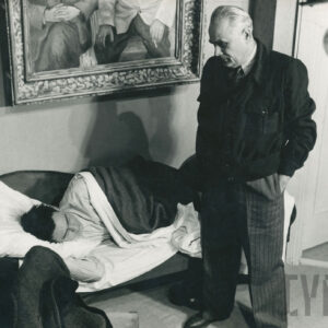 na otomanie leży mężczyzna przykryty kołdrą, obok stoi drugi mężczyzna w spodniach w pasy i marynarce, ręce ma w kieszeniach, na ścianie fragment obrazu