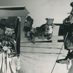 na scenie znajdują się cztery lalki, kot w długiej sukni przed domkiem, dwa kocięta na płocie i mężczyzna w płaszczu z wąsami