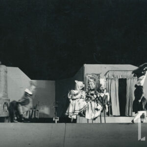 pośrodku sceny znajdują się trzy lalki - świnia, kot oraz koza w eleganckich sukniach, po ich obu stronach inne lalki, z tyłu pianino, drzwi z zasłonami i piec