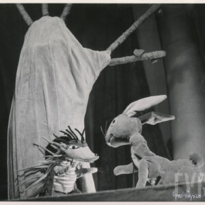 naprzeciw siebie stoją kukiełki przedstawiające lisa i zająca, za nimi dekoracja w formie drzewa