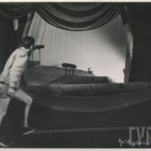 mężczyzna ubrany w szorty z lornetką w ręce, stoi zwrócony w kierunku sceny, na której znajduje się kukiełka przedstawiająca zająca